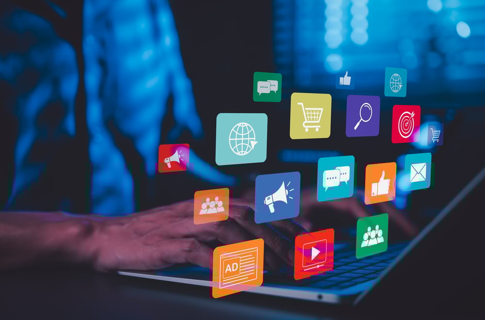 Hands typing on a laptop surrounded by floating digital marketing icons representing social media, video content, e-commerce, and online advertising. Visualizing omnichannel marketing, content repurposing, or cross-platform video strategy.