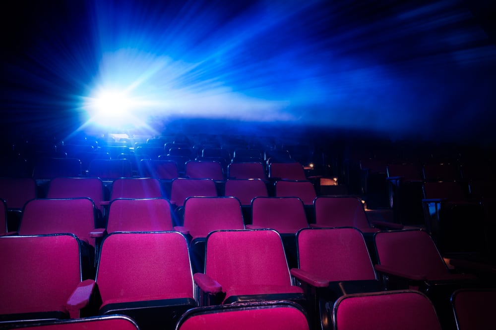 An empty movie theater with rows of red seats illuminated by a bright blue light from a projector in the back, casting dramatic beams through the haze in the air.